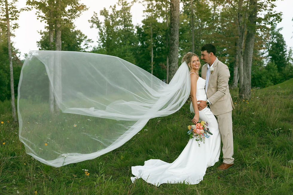 Bride and groom pose in a grassy forest at an outdoor wedding venue in West Virginia, her long veil streaming left in the wind as she smiles and holds a colorful bouquet.