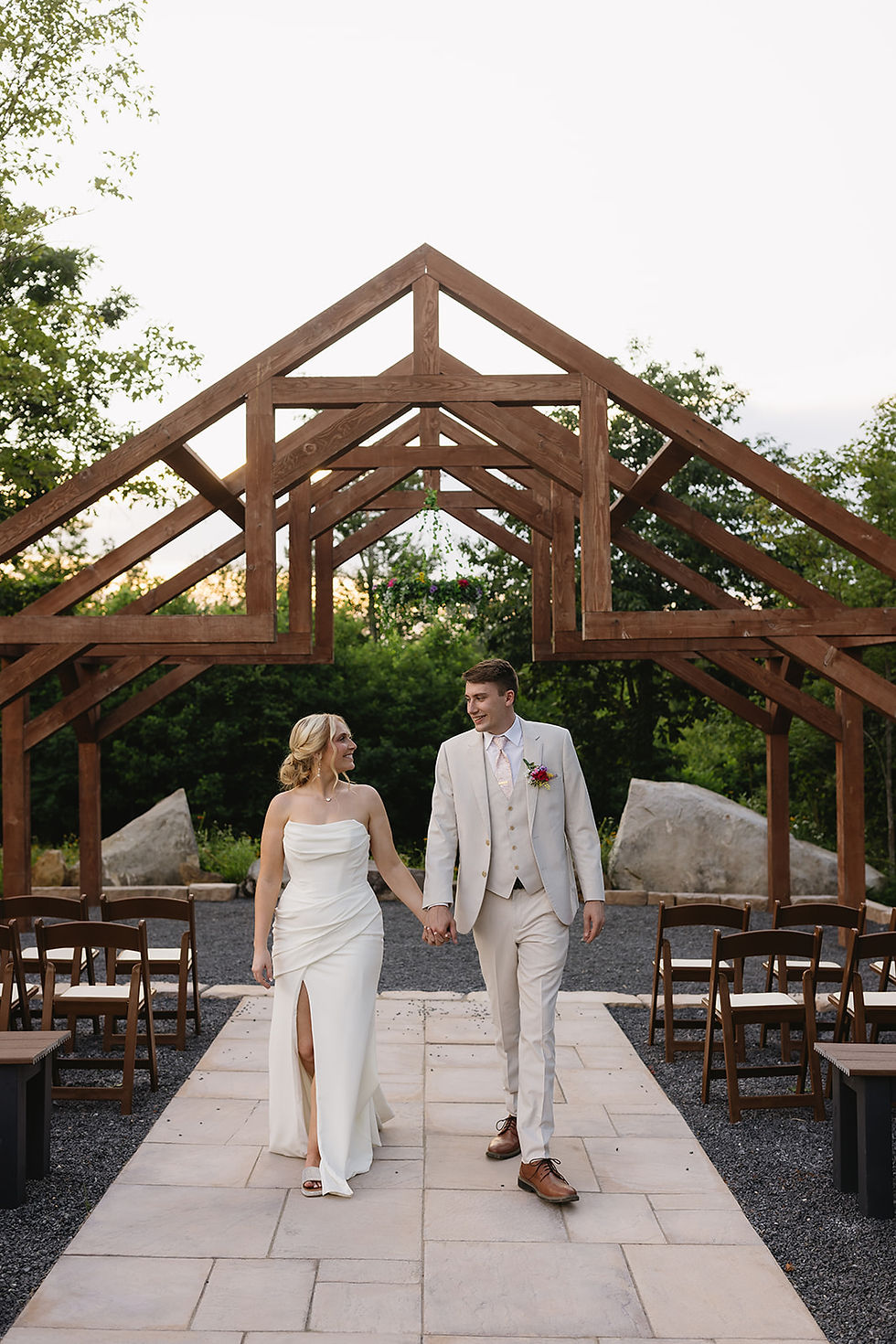 Bride and groom walk hand in hand under a wooden arch at wedding venue in Charleston, WV, smiling, in an outdoor wedding setting with greenery and chairs.