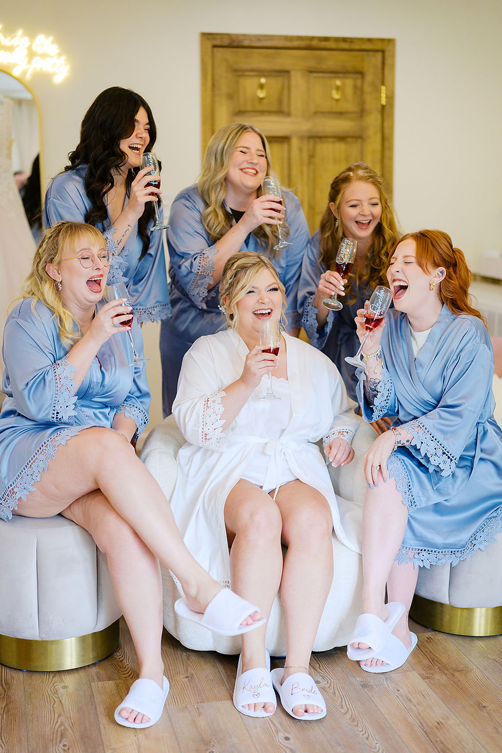 Bridesmaids in blue robes and bride in white robe laugh and raise glasses in a cozy bridal suite room. Joyful mood, with champagne flutes and wooden door in the background at wedding venue in Charleston, WV.