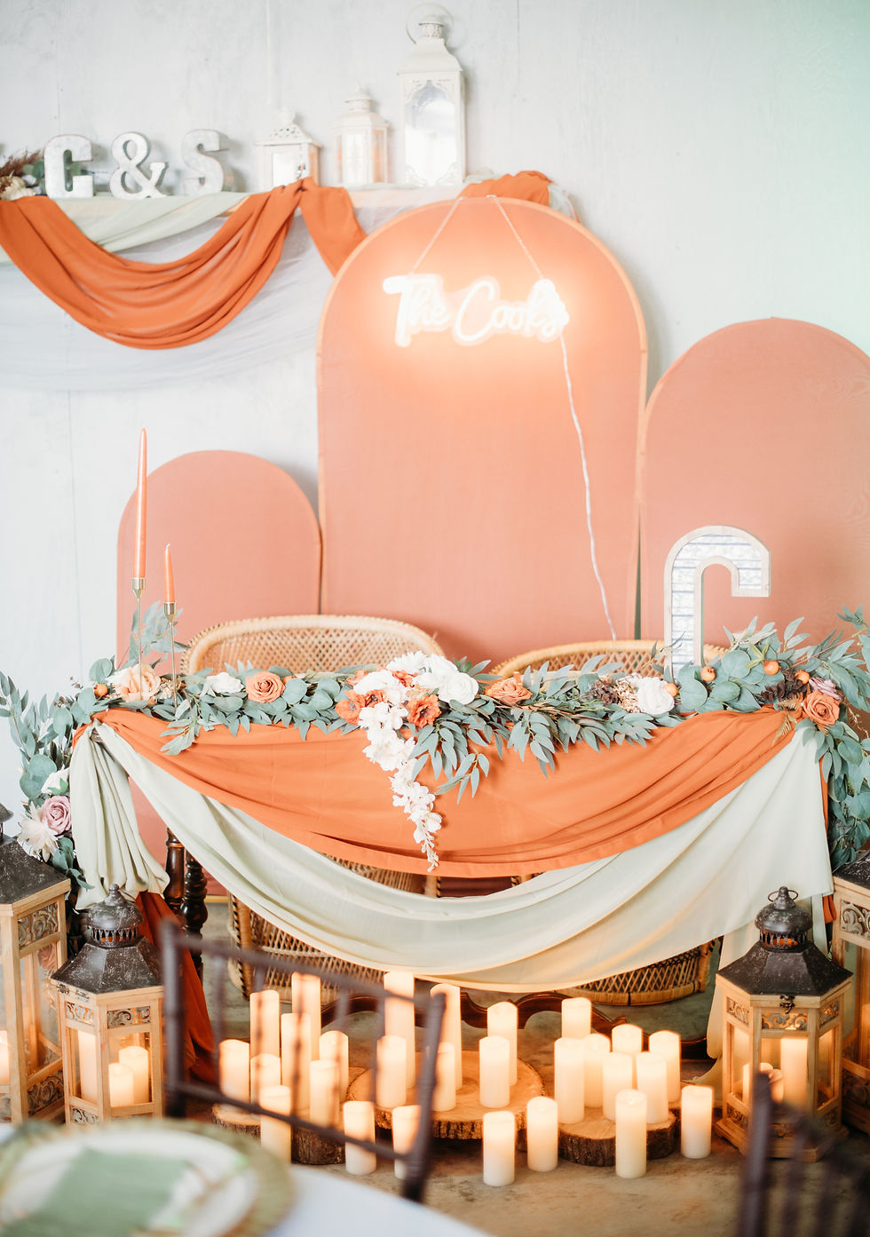 Elegant wedding setup with terracotta and sage green drapery, greenery, foral, candles, and a neon sign reading "The Cooks." Lanterns add warm ambiance at luxurious West Virginia wedding venue.