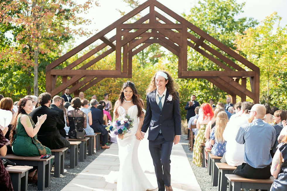 Bride and groom walk down an outdoor West Virginia wedding venue aisle, holding hands. Guests applaud under a wooden arch, surrounded by lush greenery during beautiful fall wedding near Charleston, WV.