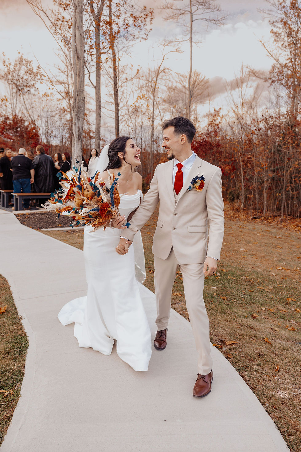 Bride and groom in beige and white attire laughing, walking on a path with a vibrant bouquet against an autumn forest backdrop.