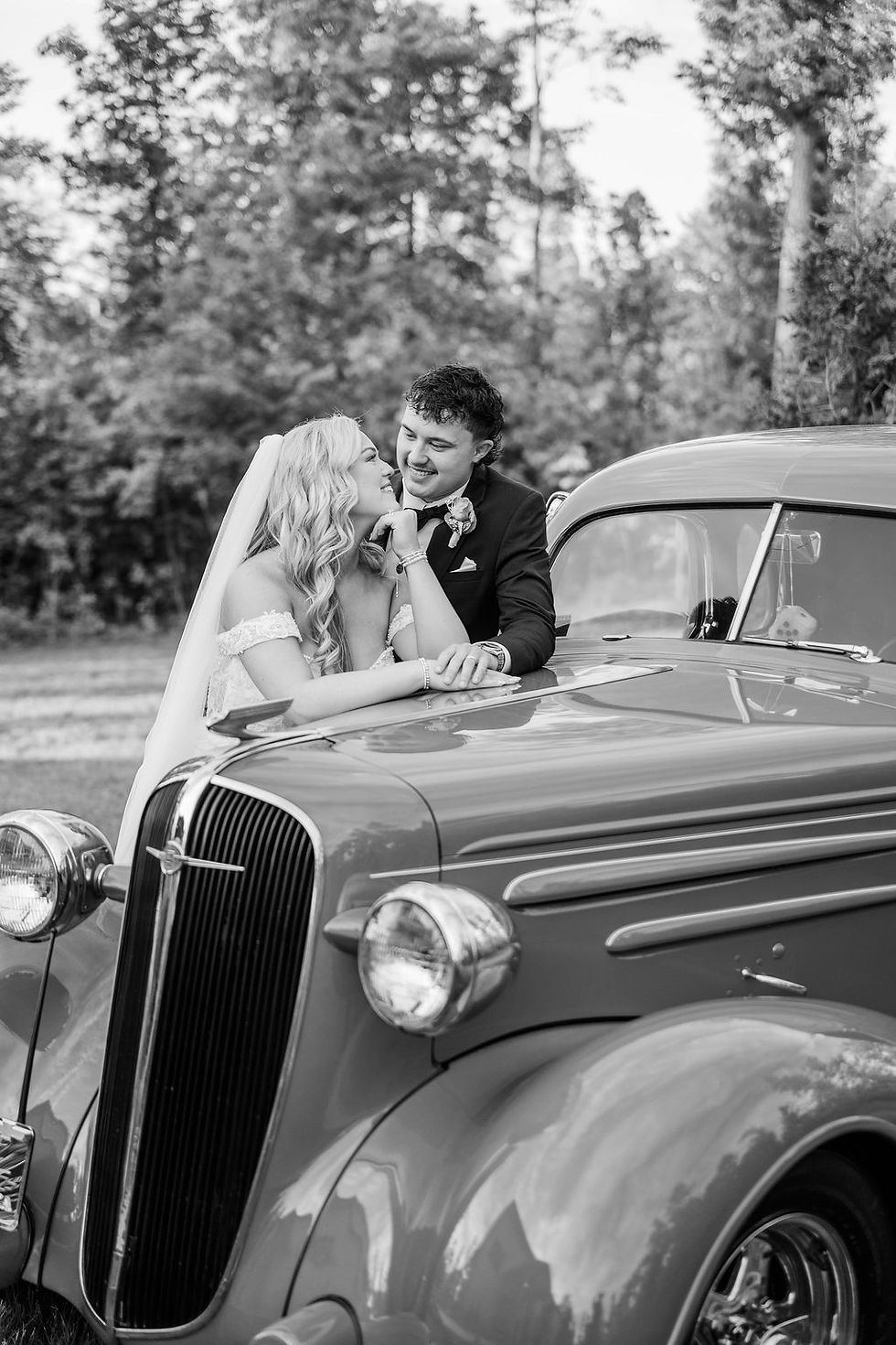 Black-and-white photo of bride and groom smile and lean on a vintage car in a wooded outdoor wedding venue in Charleston, WV.