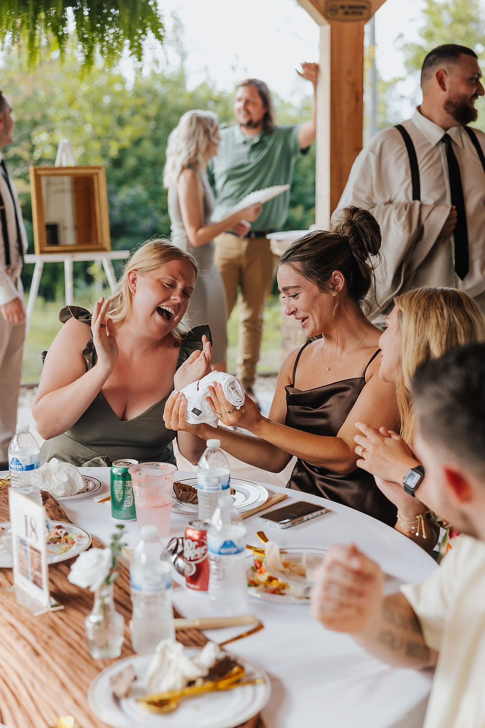Wedding guests laugh at an outdoor wedding reception venue in West Virginia while a woman unwraps a rolled napkin at a table with drinks and plates.
