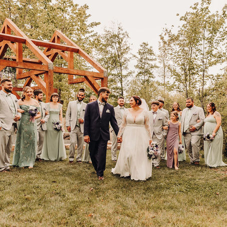 Bride and groom walk hand-in-hand with their wedding party, wearing pastel outfits, in a lush outdoor West Virginia wedding venue with a wooden open-air chapel.