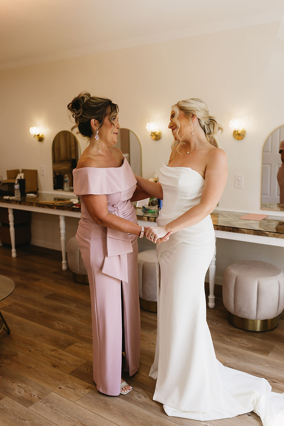Bride in fitted wedding dress and mother of bride in a soft blush mother of the bride dress smiling and holding hands in a bridal suite with wooden floors and mirrors at a Charleston, West Virginia wedding venue.