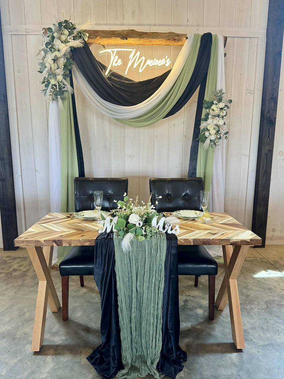 Wedding sweetheart table setup with "Mr & Mrs" sign, green and black drapes, and floral decor. A custom neon "The Macioces" sign is in the background at Charleston, West Virginia wedding venue