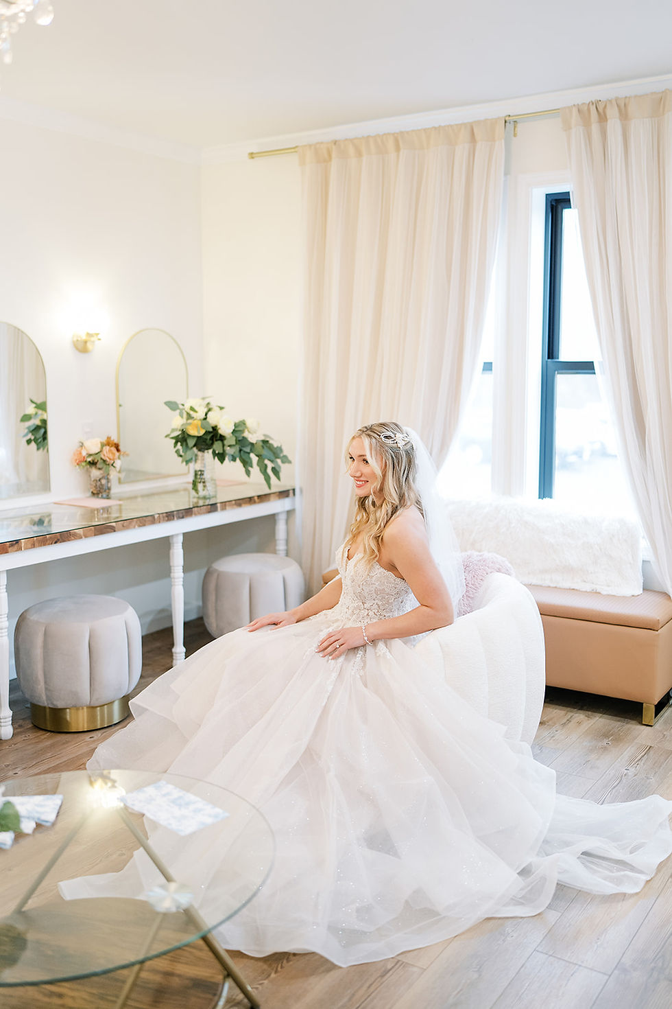 Bride in a white gown sits on white plush chair in a bright Charleston, WV wedding venue bridal suite with mirrors, flowers, and pastel decor, looking serene and smiling