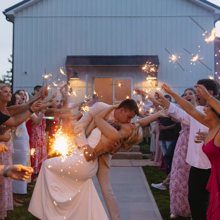 A bride and groom kisses passionately beneath sparklers during wedding exit held by guests in colorful attire outside a white reception barn in Charleston, WV, creating a festive, romantic scene.