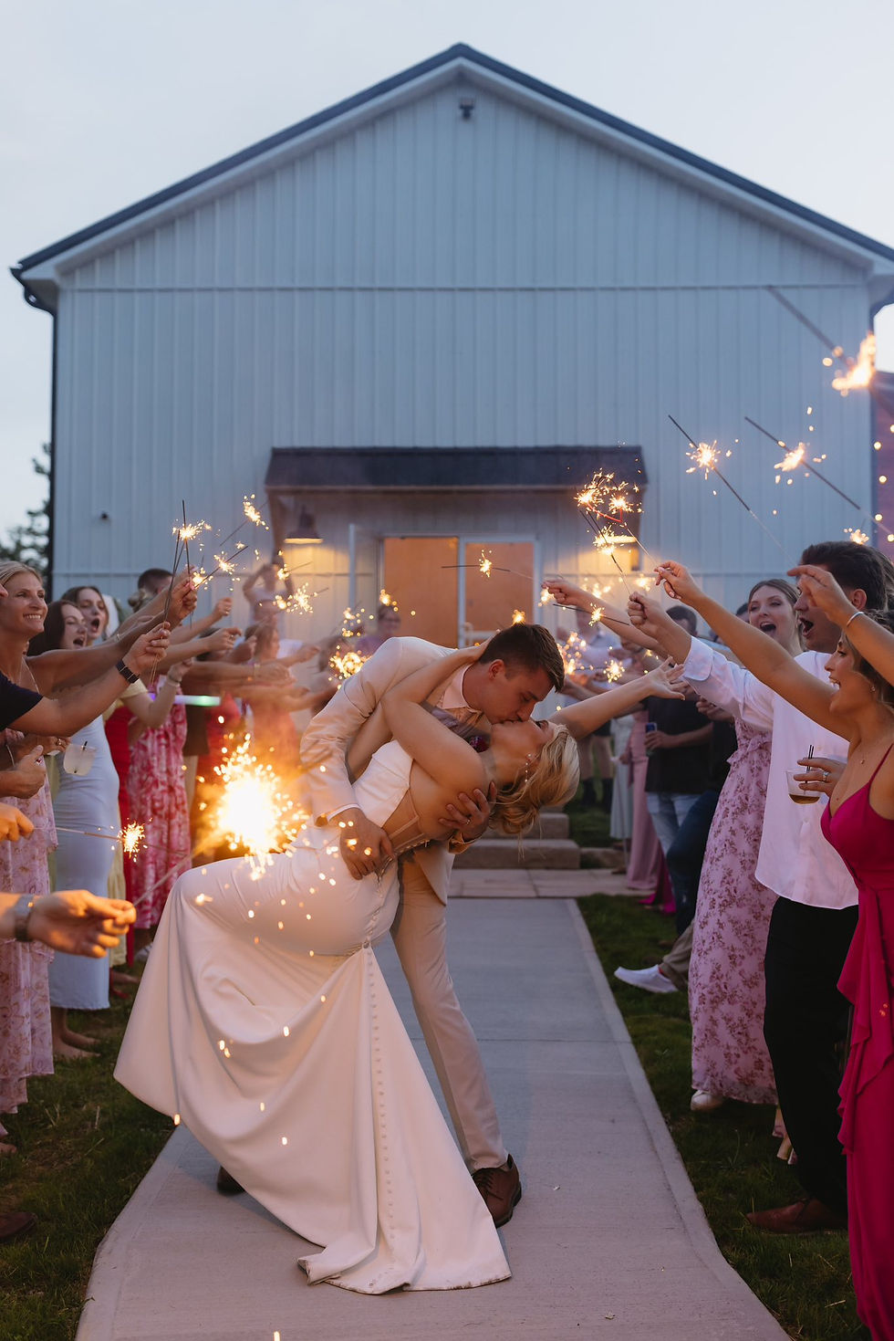 A bride and groom kisses passionately beneath sparklers during wedding exit held by guests in colorful attire outside a white reception barn in Charleston, WV, creating a festive, romantic scene.