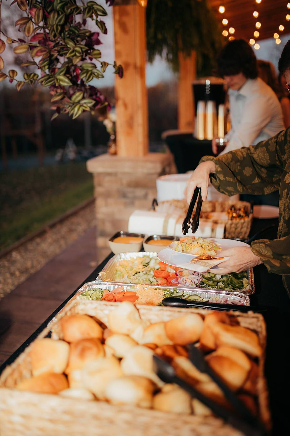 Person serving food at a buffet with salads, rolls, and tongs. Outdoor reception venues in West Virginia with string lights, wood and stone pillars, and greenery.