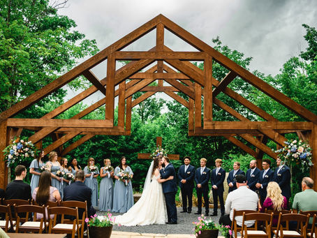 Bride and groom kiss at an outdoor wedding location in WV under a wooden arch, with bridesmaids, groomsmen, and guests seated nearby.