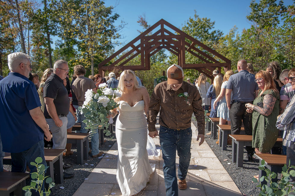 Bride and groom joyfully walk down the aisle at outdoor West Virginia wedding venue under a wooden arch. Bride holds white flowers, guests watch, and bird seed floats around them.