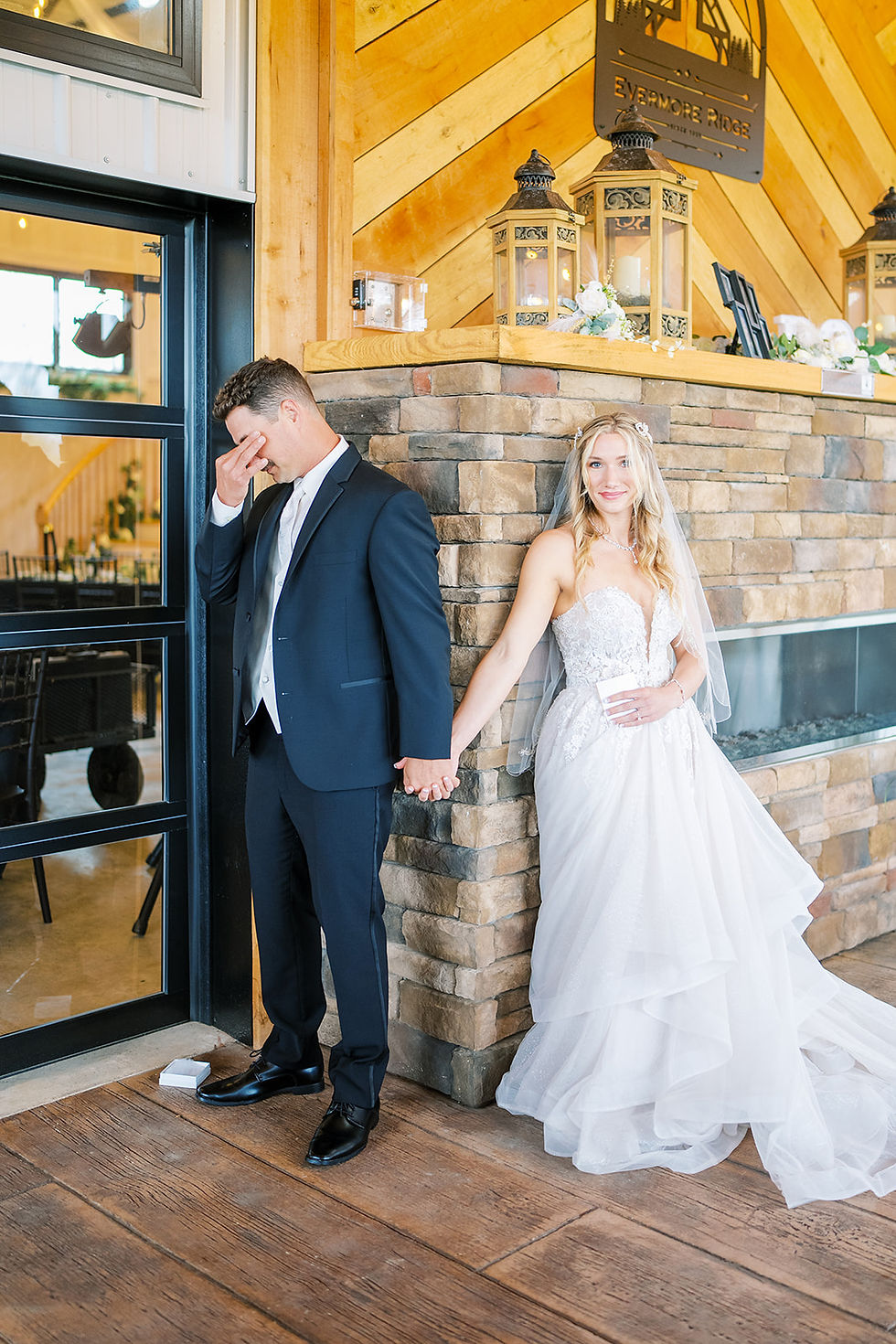 Bride and groom hold hands during a first touch at a wedding venue in Charleston, WV. Around a stone fireplace, not seeing each other. Bride smiles in white wedding dress, groom in black suit wipes tears from his eyes.