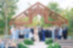 A group of people poses happily under a wooden arch in an outdoor West Virginia wedding location, surrounded by greenery. Many wear blue with aisle lined by lush green ferns.