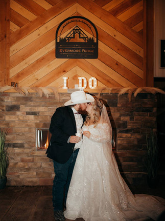 stone fireplace with words "I DO" glowing on mantle. Black Evermore Ridge wedding venue sign hangs above. Bride in long sleeve lace gown kisses groom in cowboy hat. Wedding venue near Charleston, West Virginia. 