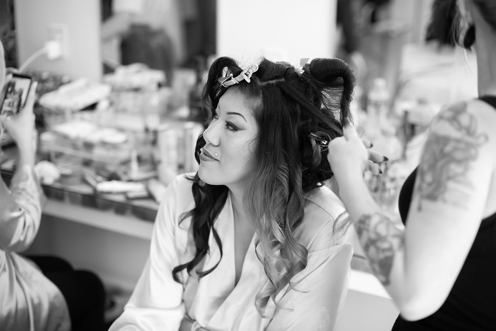 Bride with curled hair being styled in a luxurious West Virginia wedding venue bridal suite. Another person uses clips, surrounded by products. Calm atmosphere, monochrome setting.