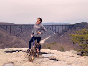 Woman with a dog stands on a rocky overlook, smiling, with the New River Gorge National Park Bridge behind her located near a New River Gorge wedding venue. 