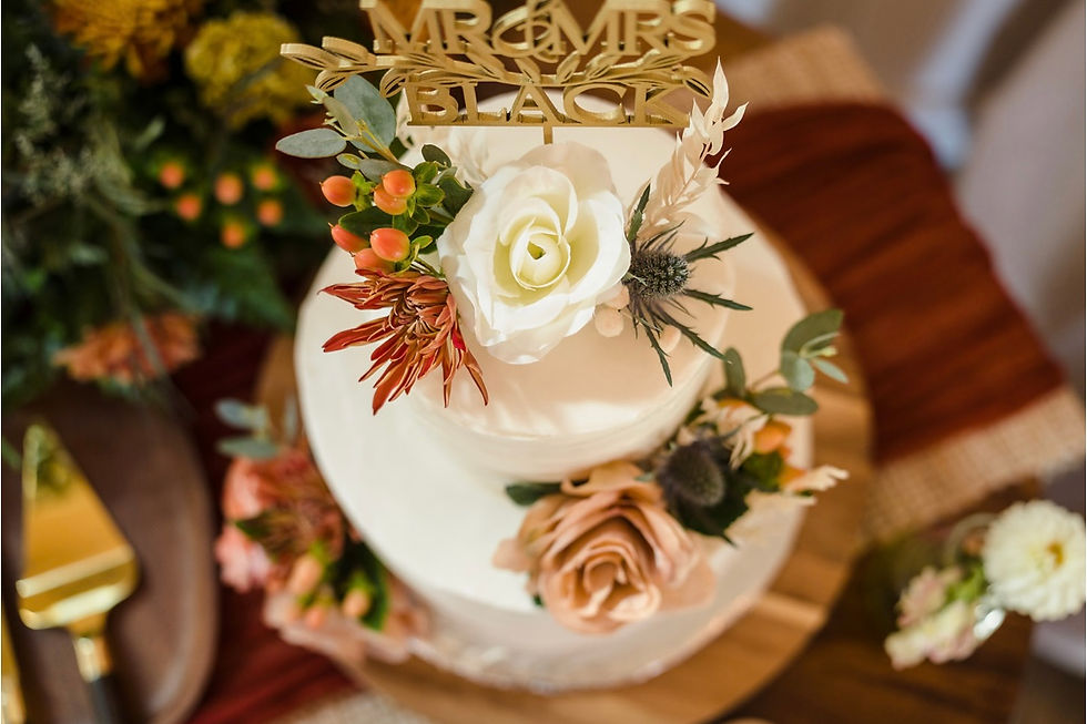 Tiered wedding cake with floral decor, "MR & MRS BLACK" cake topper. Warm, neutral tones, gold cake server on rustic table with blurred floral backdrop at luxurious West Virginia wedding venue