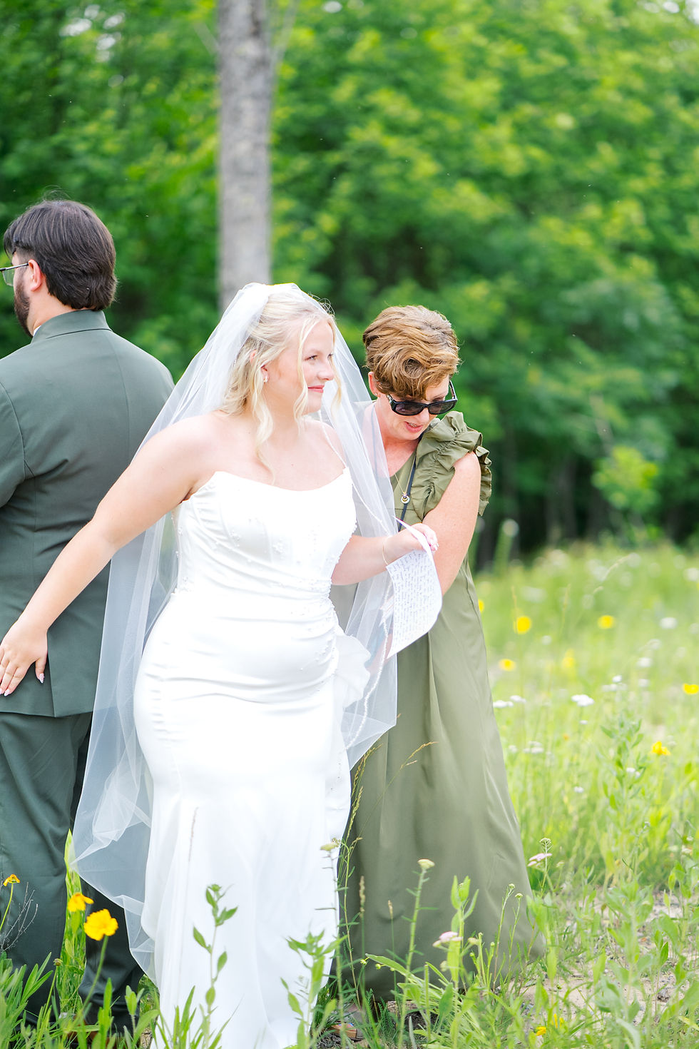 Bride in white dress holds a note, smiling with her day of coordinator in green dress, Imagine Designs by Beck, getting the couple ready for their private vows. In a field of wildflowers at luxurious West Virginia wedding venue.