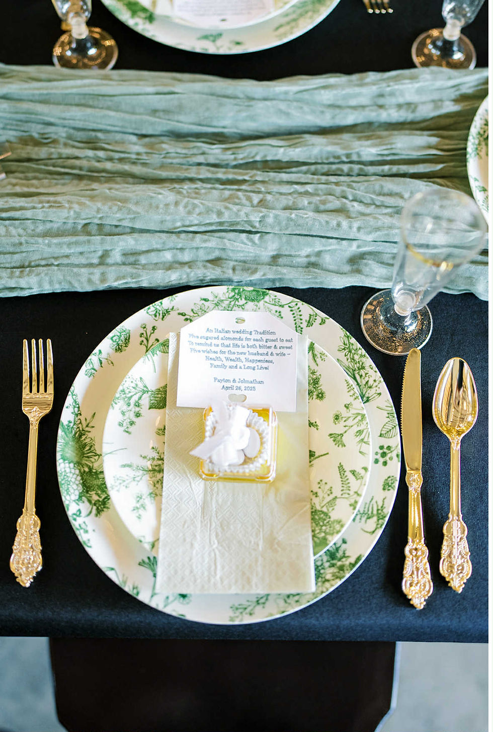 Elegant table setting with floral plates, gold utensils, a menu card, and clear glasses on a textured green runner with a unique wedding favor idea featuring groom's Italian heritage at luxurious West Virginia wedding venue