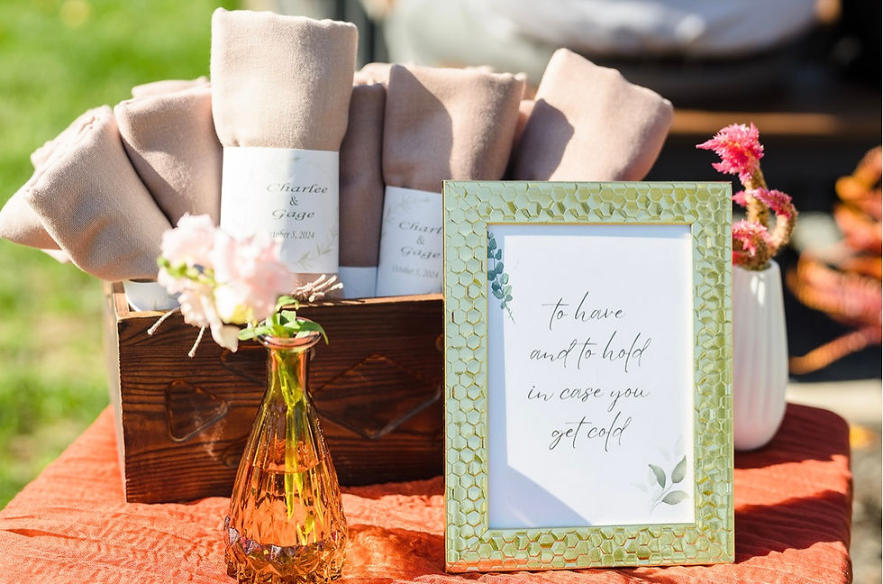 Framed sign reads "to have and to hold in case you get cold" beside pink flowers, beige blankets, and rustic box on rust tablecloth at outdoor ceremony site at Charleston West Virginia wedding venue