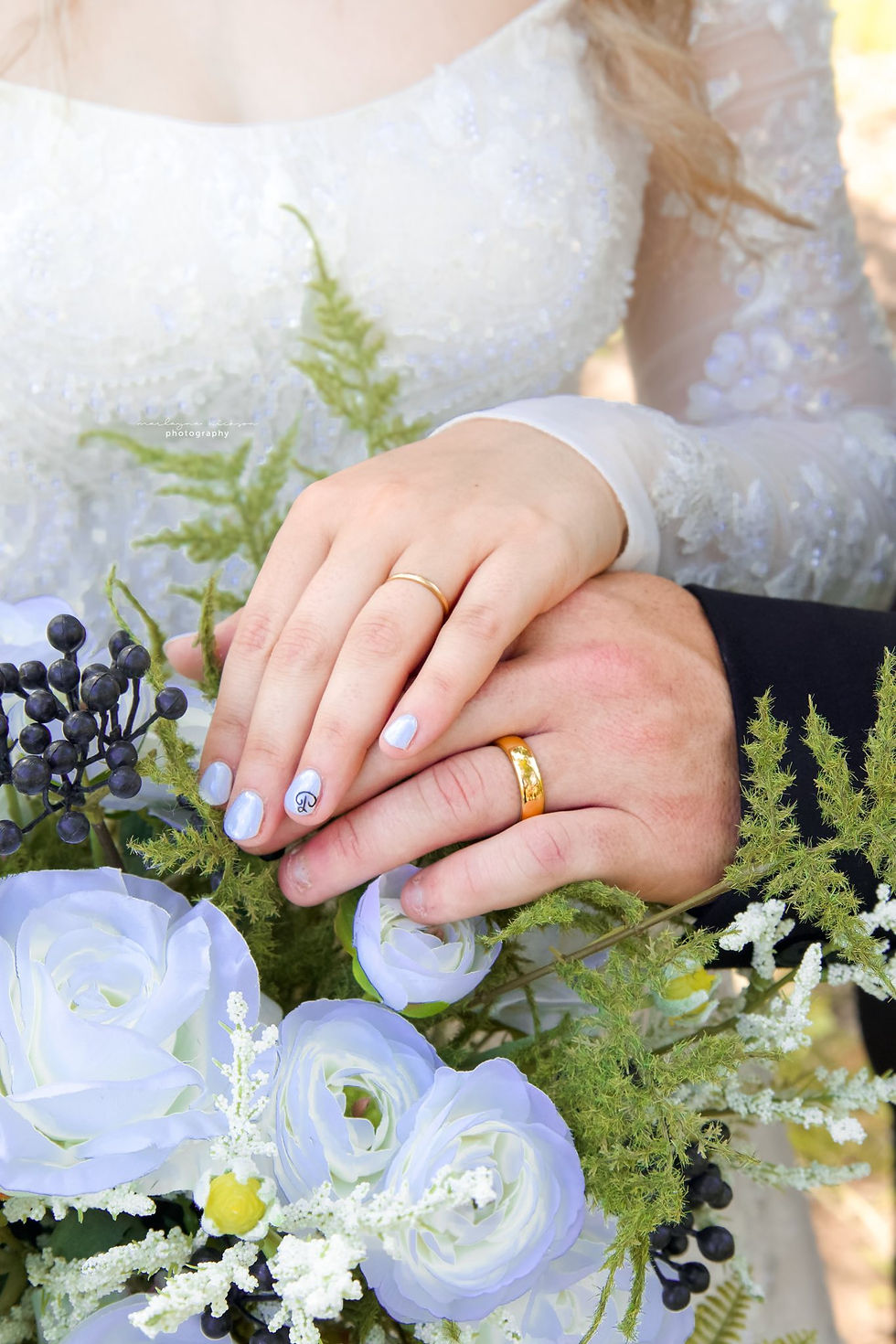 Bride and groom's hands with gold wedding rings rest on light blue and white roses with greenery. Bride's nails are white with initials on ring finger at luxurious West Virginia wedding venue.