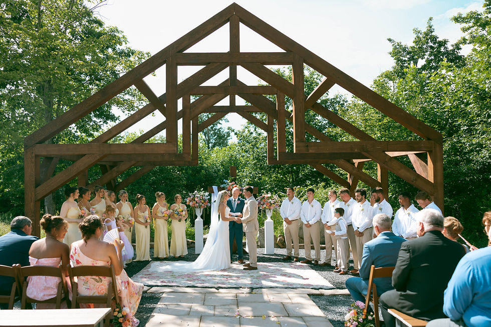 Outdoor wedding venue in West Virginia under a wooden open air chapel, bride and groom exchanging vows with bridal party and guests seated amid lush trees.