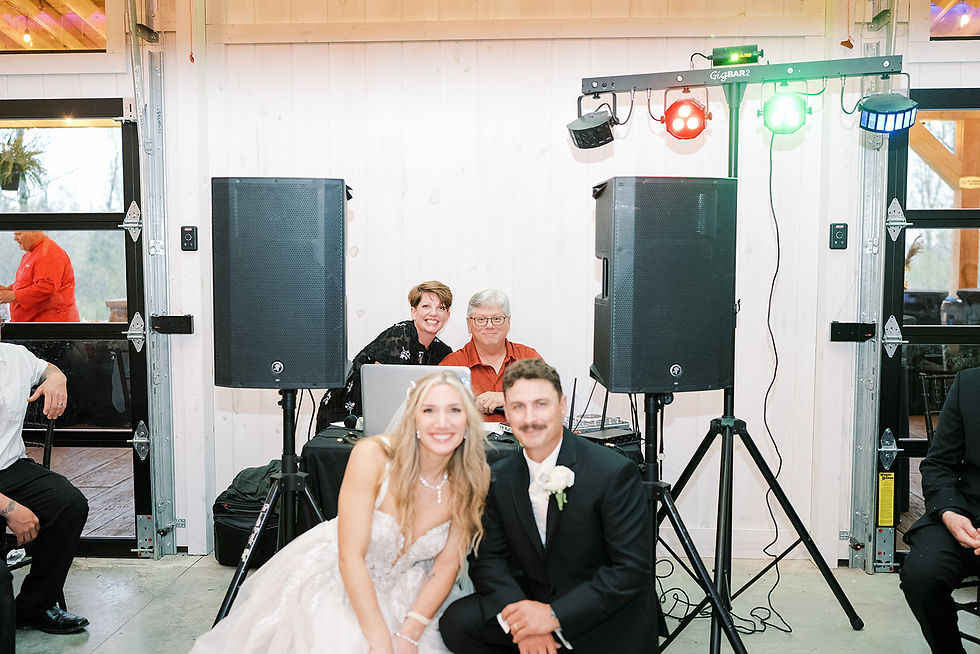 Bride and groom smile in front of DJs with sound equipment. Bright lights and white wooden walls create a festive atmosphere at Charleston, WV wedding venue.