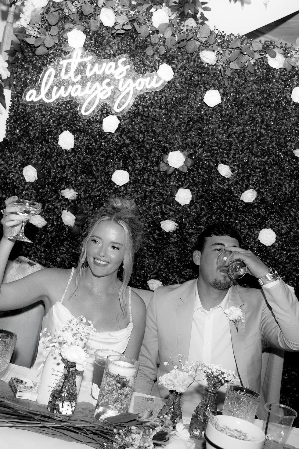 Bride and groom toast at a floral wedding backdrop at an outdoor wedding venue in West Virginia with neon sign It was always you, smiling amid roses and table flowers
