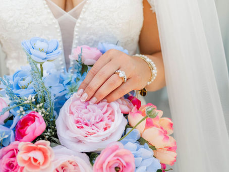Bride in a white beaded wedding dress and veil holds a pastel pink, blue, and peach bouquet, showing her engagement ring and nails at a wedding venue in Charleston, WV.