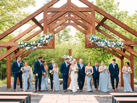 Wedding party in blue and navy attire under a wooden outdoor arch with floral decor, joyful mood, outdoors with green trees in the background at outdoor ceremony site in WV.