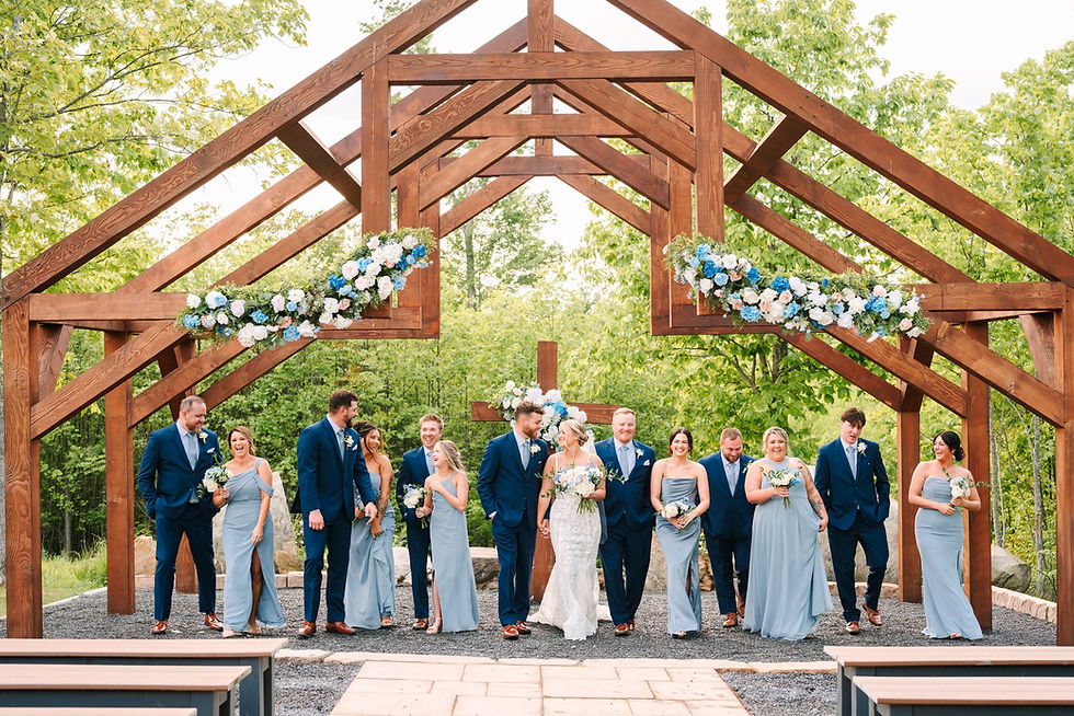 Wedding party in blue and navy attire under a wooden outdoor arch with floral decor, joyful mood, outdoors with green trees in the background at outdoor ceremony site in WV. 