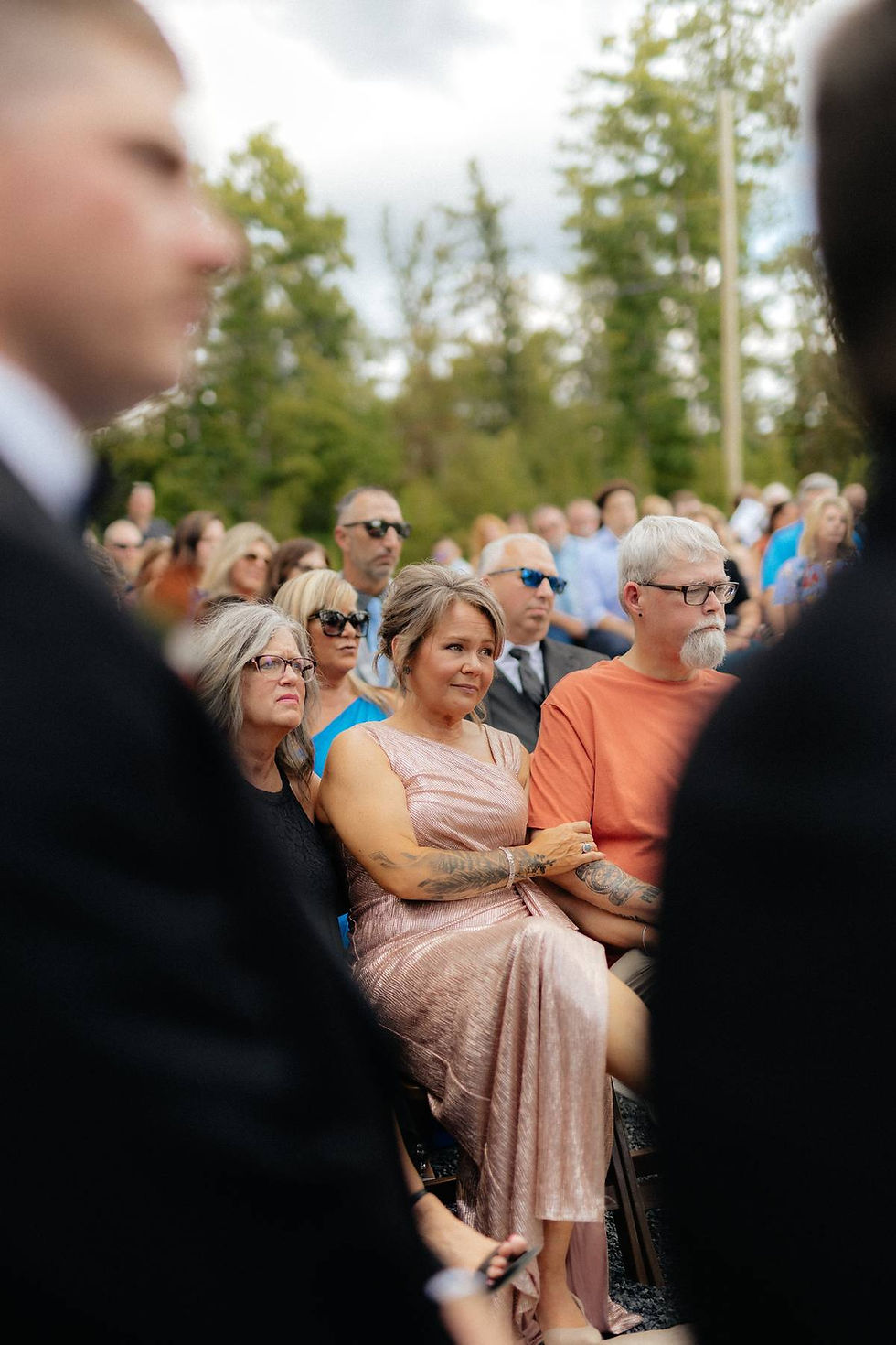 Wedding guests sit at outdoor wedding location in WV. The mother of the groom in a pink dress looks ahead watching her son get married, her hand on a man's arm. 