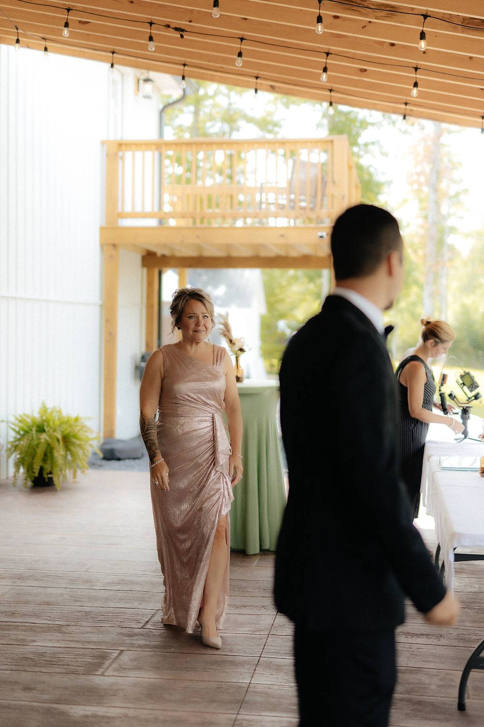 Mother of the groom in a shimmering pink dress walks under a wooden patio with string lights at a Charleston, West Virginia wedding venue. A groom is in the background waiting for his mother to approach him on his wedding day. 