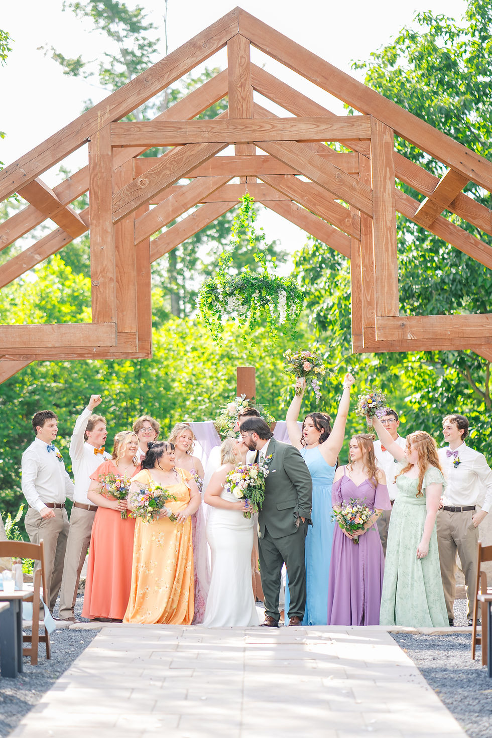 Wedding party at outdoor wedding venue in West Virginia with wooden arch, bride and groom kiss, bridesmaids in colorful dresses, groomsmen in white shirts, joyful mood.