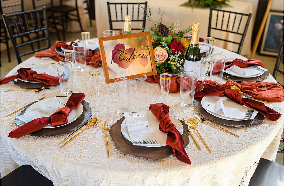 Elegant table setting with a lace cloth, beige tablecloth, gold cutlery, terracotta napkins, and champagne glasses. A table number sign reads "Thirteen." Floral centerpiece at luxurious West Virginia wedding venue.