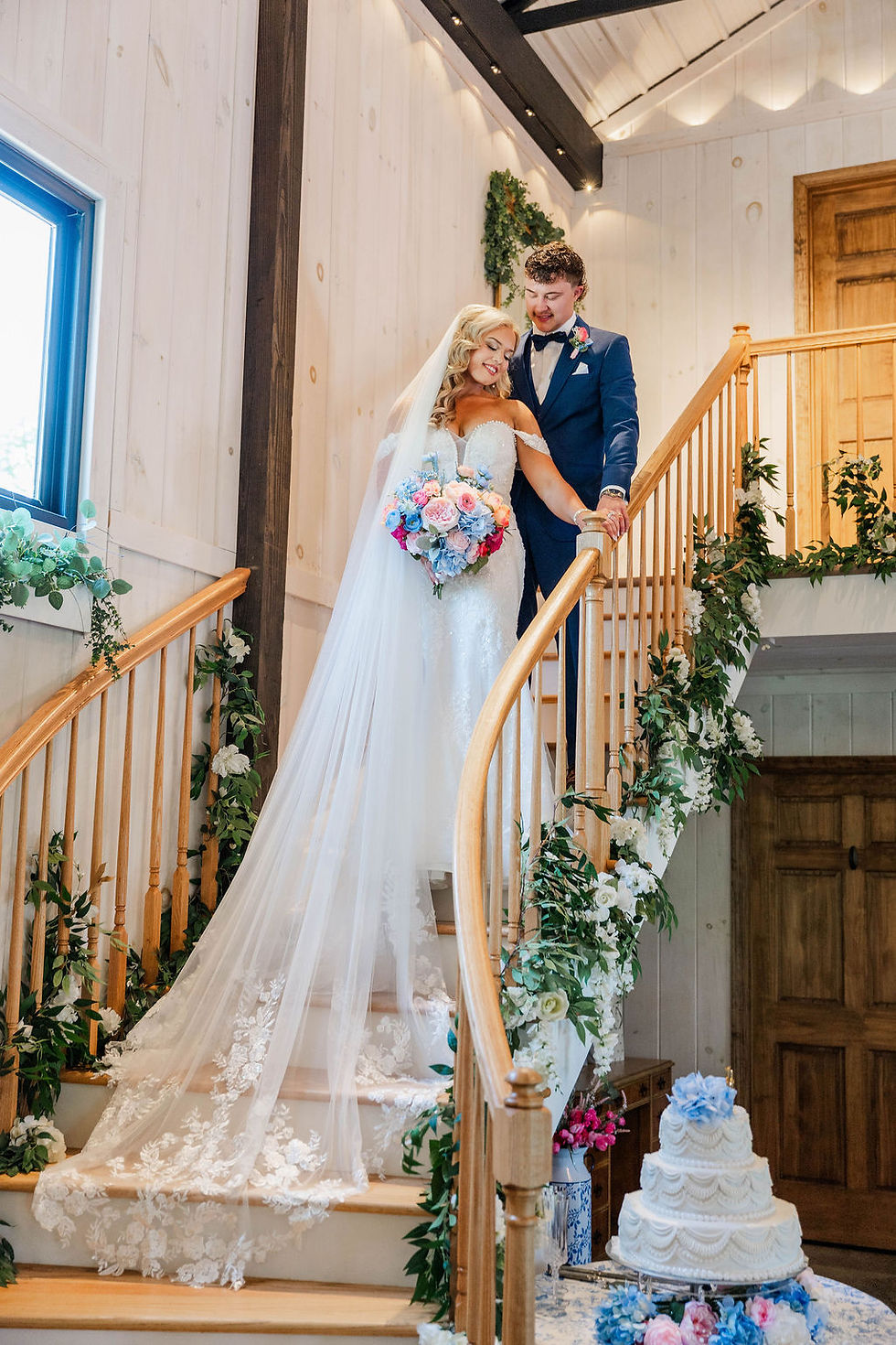 Bride and groom in wedding attire descend a flower-lined staircase at a wedding venue in Charleston, WV, with a white tiered cake below.