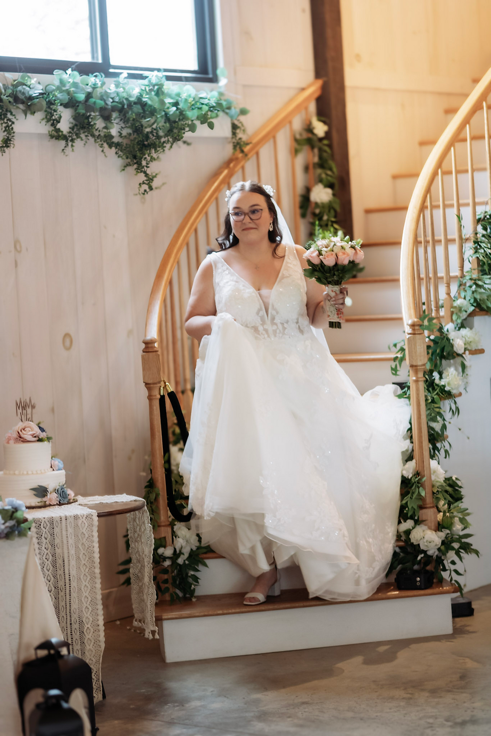 Bride in a white wedding gown descends a flower-adorned wooden staircase inside a reception venue in West Virginia, holding pink roses. Cake with "Mr & Mrs" topper on a table nearby.