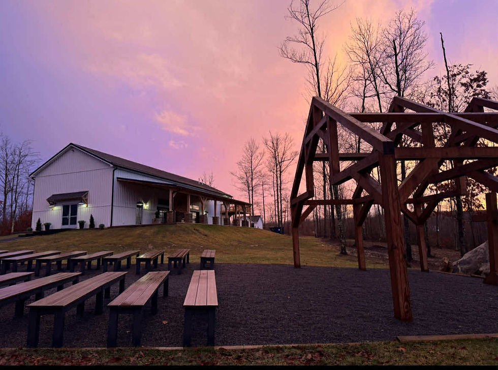 Outdoor wedding venue near Charleston West Virginia at sunset. Rustic wooden arch, benches, white barn on a grassy hill. Pink-orange sky with bare trees. Peaceful mood.