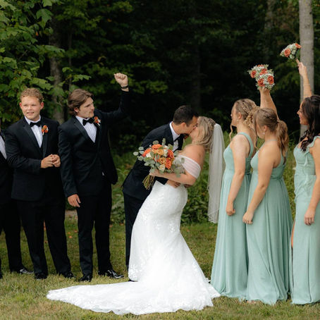 Bride and groom kiss at outdoor wedding venue in West Virginia, surrounded by groomsmen in black suits and bridesmaids in sage green dresses, with greenery in the background.