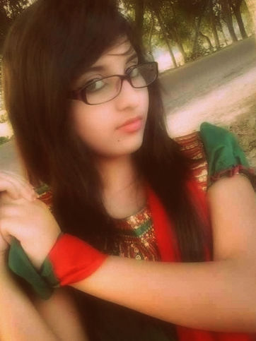 A young woman with glasses, wearing a colorful traditional outfit, posing outdoors with a red scarf tied around her arm and trees in the background.