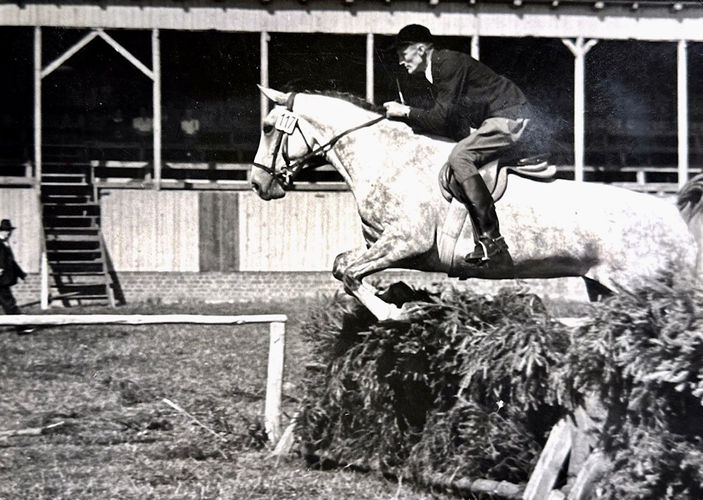 Eine alte schwarzweiß Aufnahme von einem Schimmel, der geritten über einen Busch springt.