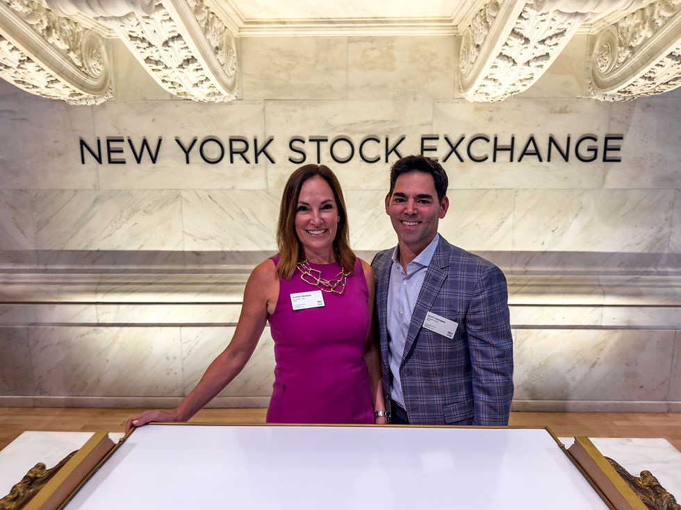 A woman in a pink dress and a man in a plaid jacket stand beneath the New York Stock Exchange sign.
