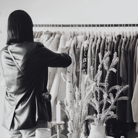 A woman browsing a rack of vintage and second-hand clothing in a boutique-style shop. She is viewed from behind while looking through curated pieces displayed on hangers.