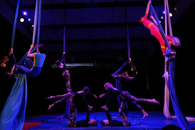 6 students perform aerial silks while four students perform partner acrobatics in the center of the stage. 