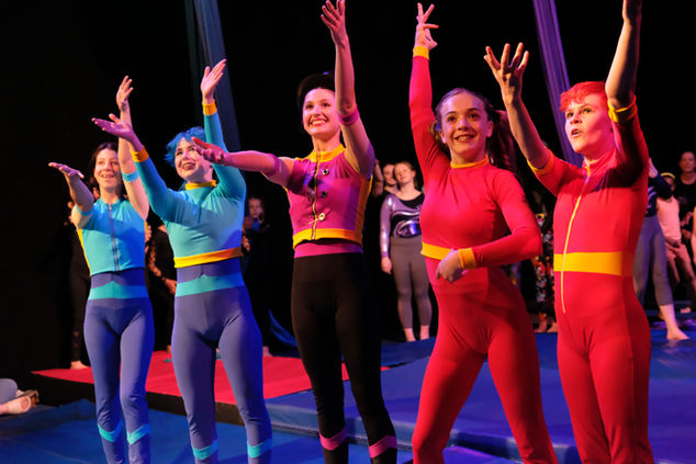 5 female bodied teenage aerial students at the end of the show stand before the audience and thank the lighting and sound team by gesturing upwards with their arms while standing in a line. 