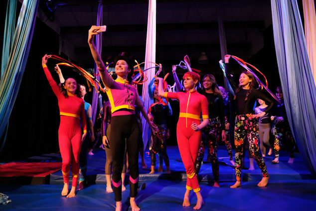A female bodied student holds up a camera towards a large group of students behind her to take a selfie of herself with the cast of the show. 