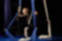 Two female bodied aerial students perform the splits on aerial silks. The student in front is white and looks at her hands, while the African American student next to her smiles at the audience.