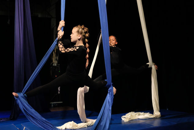 Two young female bodied students perhaps age 8-10 perform the splits on aerial silks. The rear student, an African American, smiles at the camera, while the white student in front looks at her aerial silks. 
